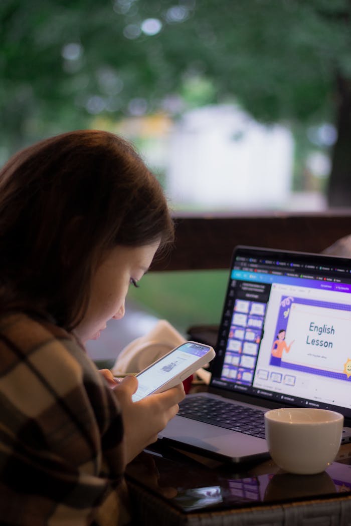 A woman is using a smartphone and laptop for an online English lesson outdoors.